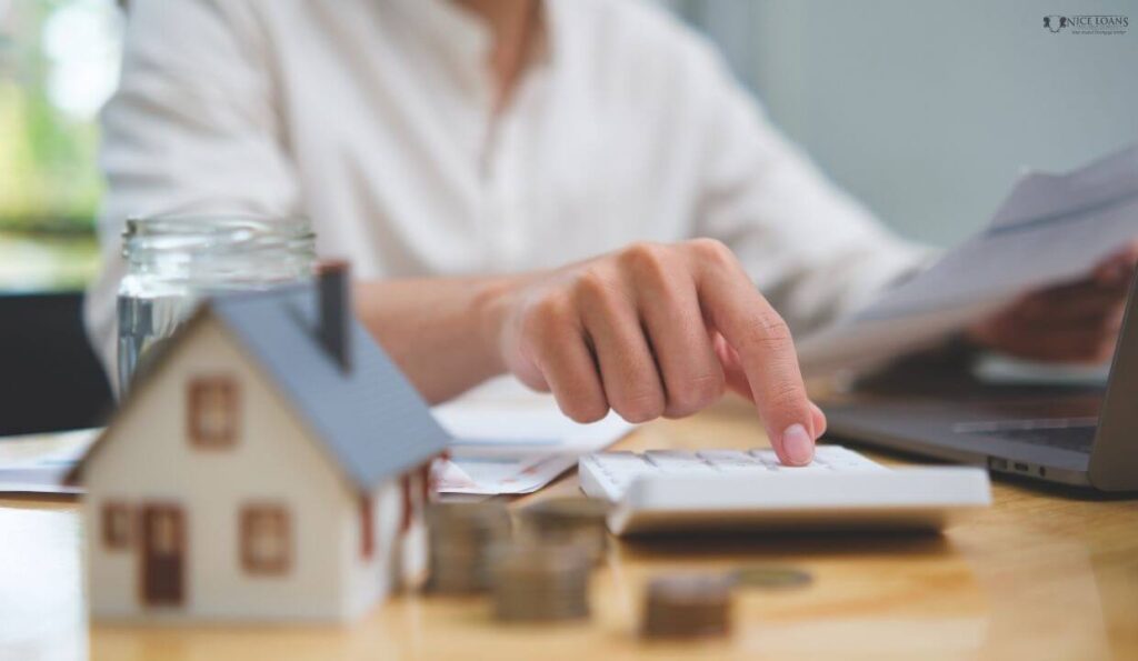 a man at his table working with a calculator, a small model home and some stacked coins are also on the same desk.