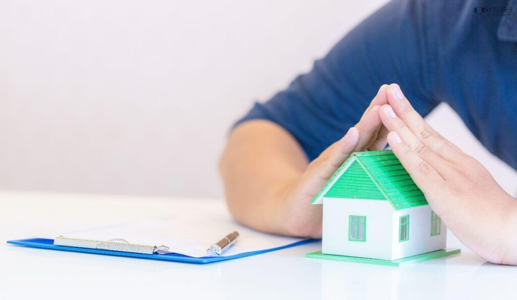 a man with his palms over the roof of a model house with a document beside it on the same table. 