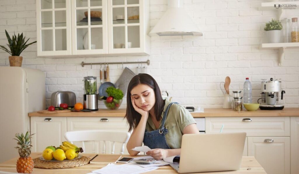 a woman with her hand on her cheek and elbow on the table with a laptop open, staring at a receipt in distress. 