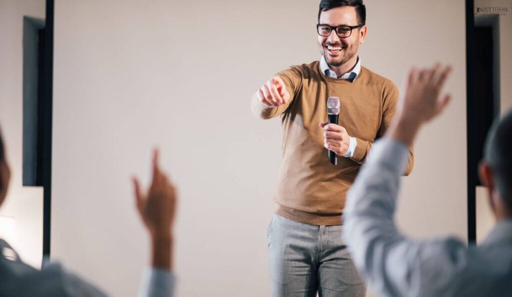 a man on a podium smiling with a mic with two people raising their hands from the audience. 
