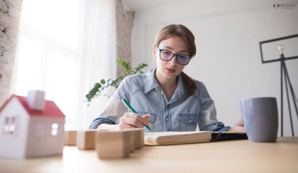 A woman at her table with a model house, a few blocks and a cup on top, working intently. 