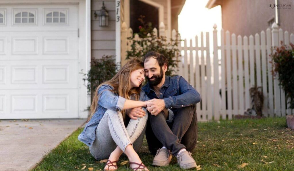 a couple outside their new home, seated on the grass, hugging. 
